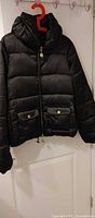 Front view of black quilted puffer jacket on hanger showing pockets, zipper, and hood