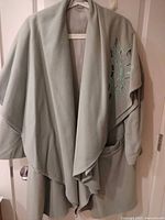 Front view of sage green loose overcoat showing open front, draped collar, pocket and geometric applique