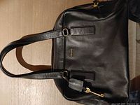 Front view showing Fossil black handbag, gold-tone zipper, twin handles, luggage tag charm