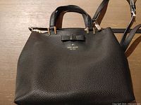 Front of black Kate Spade pebbled leather handbag showing bow detail, top handles and strap