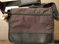 Front view of Roots messenger bag showing flap, ROOTS logo, front zipper pocket, attached shoulder strap and plush bear keychain