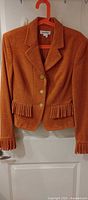 Front view of orange blazer on hanger showing three buttons, lapel, pleated fringe trim at waist and cuffs