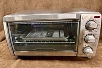 Front view of toaster oven showing stainless steel finish, glass door, control panel, interior with manual on rack