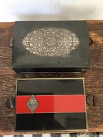 Two metal boxes on wood surface; larger floral-inlaid box above smaller red-band box