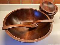 Large maple serving bowl with wooden fork and spoon resting across, stack of small bowls at rear