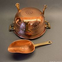 Copper colander upside down showing perforated base, brass handles, and scoop front view