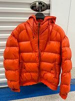 Front view of hooded orange puffer jacket showing zipper, pockets, hood and ribbed hem