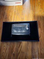 Front view of black leather wallet showing exterior zip pocket and attached charm