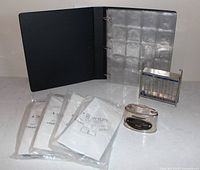 Lot overview showing binder with pocket sheets, four packs of coin flips, clear acrylic coin bank, metal coin bank