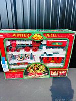Front view of boxed Winter Belle Express train set showing locomotive, freight car, caboose and holiday scene