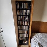 Full view of tall black media bookcase filled with CDs beside closet and bed