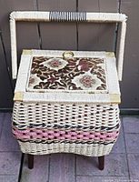 Front view of wicker sewing basket showing floral tapestry lid, brass corners, woven handle and wood legs
