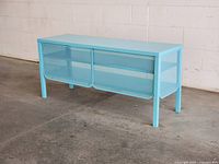 Front angled view of teal metal TV stand showing sliding mesh doors and shelves