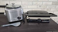 Cuisinart rice cooker with lid and paddle beside Oster panini grill