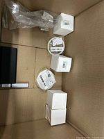 View of assorted sealed items in cardboard box, showing white cube boxes, rolls, black component