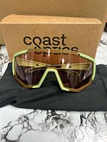 Front view of moss green frame sunglasses with pink crystal lens on microfiber pouch in front of Coast Optics box