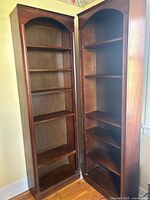 Two tall matching wooden bookcases side by side, showing interior shelves and arched tops