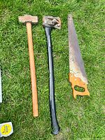 All three tools laid out on grass: splitting axe, sledge hammer, hand saw