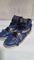 Three-quarter view showing blue leather sneakers and gold MOSCHINO logo straps