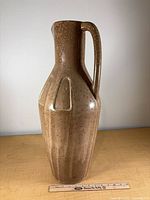 Full front view of tall beige mottled ceramic ewer with handle beside ruler