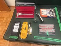 All four items laid out on green bench: caliper in red case, flaring tool box, ignition module case, leak seeker with probe and black case