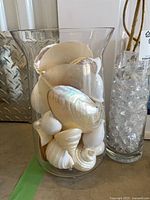 Glass vase filled with seashells beside narrow vase with clear filler