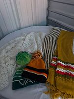 Full view of two white crochet scarves, brown/cream scarf, mustard stripe scarf, green and orange pom-pom beanies