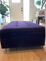 Front view of deep purple upholstered ottoman showing tufted top and brass-tone legs
