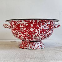 Side view showing red and white splatter enamel colander, handle, black rim, pedestal base