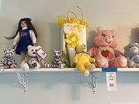 Overall view of first section of shelf showing porcelain doll, tie-dye bears, white bear, baby Pooh, pink and blue Care Bears