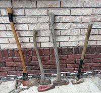 Full view of four handled tools and loose head leaning against brick wall