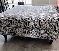 Front angled view of square ottoman displaying snow-leopard upholstery and wooden legs