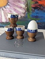 Four ceramic and wood Blue Willow egg cups, one holding an egg, shown on book