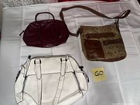 Image shows three handbags: burgundy faux-croc top-handle, tan patterned crossbody with brown trim and pockets, white faux-leather handbag with dual handles