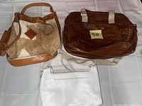 Three handbags laid out: cowhide bucket, brown tote, white crossbody