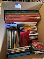 Open box showing four large hardcover books, stacked Olympic booklets, bag of lanyards