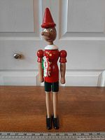 Full view of entire wooden Pinocchio figure standing next to ruler for scale