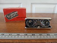 Opera glasses open beside original box on ruler showing size