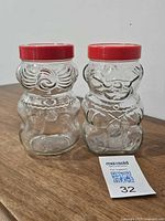 Front view of two clear glass bear jars with knitting motif and red lids