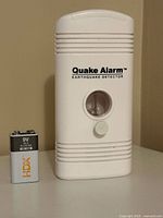 Front view of Quake Alarm detector next to HDX 9V battery