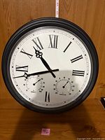 Front view of round wall clock showing Roman numerals, thermometer and hygrometer sub-dials