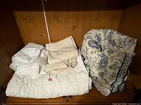 White synthetic comforter, beige fitted and flat sheets, white mattress pad, blue floral duvet displayed on shelf