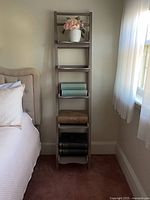 Full view of tall narrow grey bookshelf beside bed