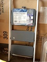Front view of folded COSCO 3-step ladder showing three grey steps and original label