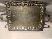 Front view of ornate silver-tone metal serving tray showing rim, handles, and engraved surface