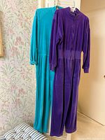 Front view of two velour jumpsuits in teal and purple hanging, showing full length and design details