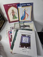 Stack of assorted vintage cross-stitch pattern leaflets visible titles The Angels’ Prayer, Elizabethan Angel, Watercolour Easter Eggs