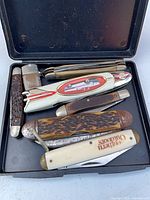 Open case displaying six pocket knives with varied handles and rusted blades