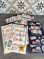 Multiple unopened sticker books showing School Sticker Book and Good Vibes cover designs