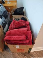 Three boxes containing red plush blankets, black faux-fur pillow and other textiles